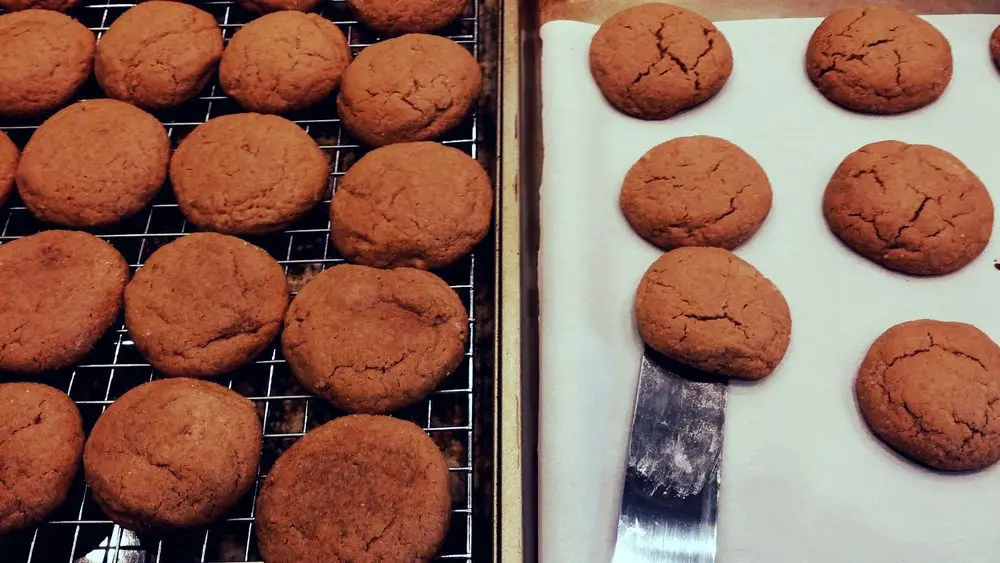 Trays of ginger cookies cooling on a rack beside a parchment-lined sheet pan