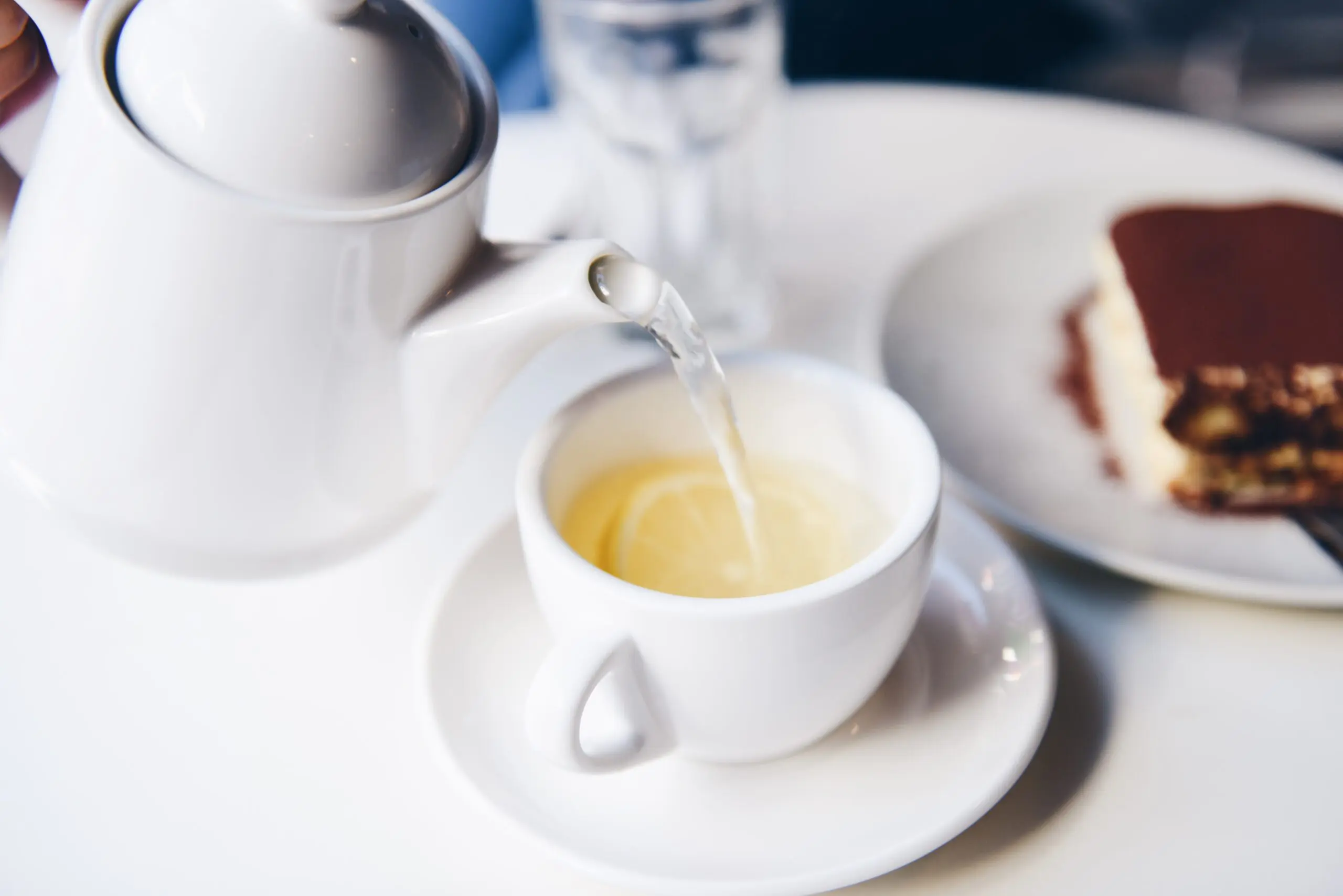 A white porcelain teapot pouring clear tea into a small cup, a slice of cake behind
