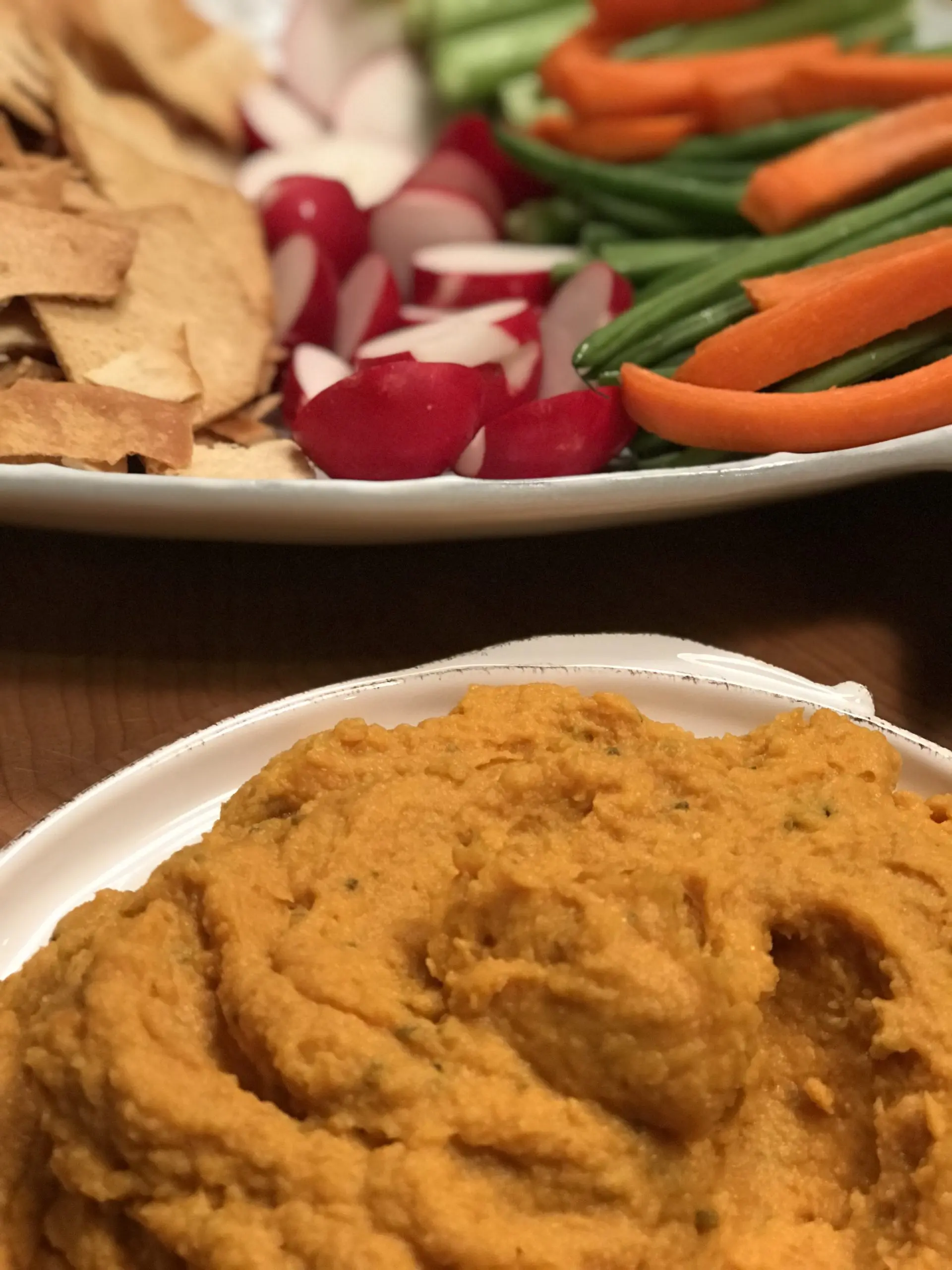 A bowl of dip with pita chips, radishes, carrots and green beans behind it