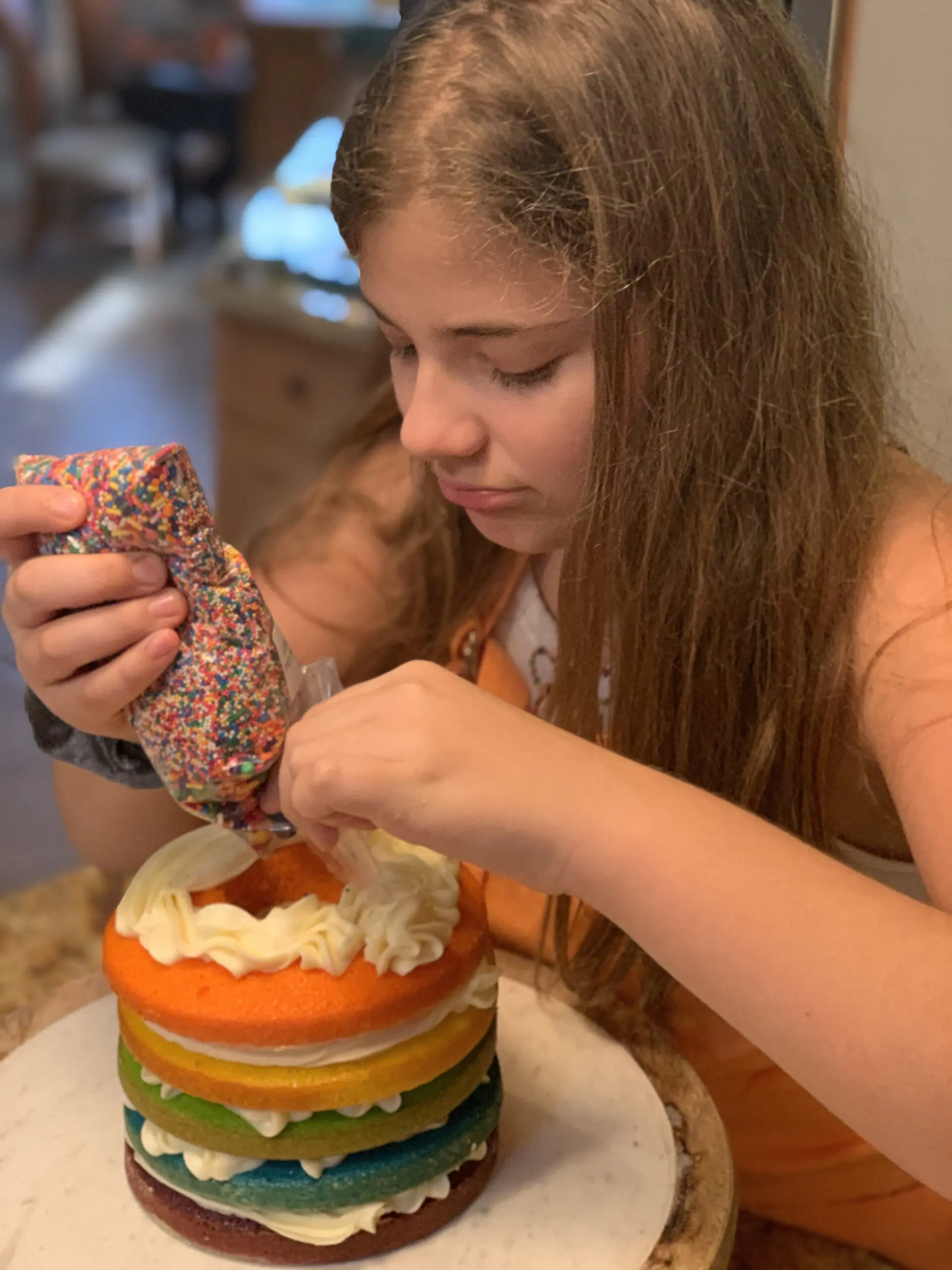 A young girl piping white frosting from a sprinkle-covered bag onto a rainbow layer cake