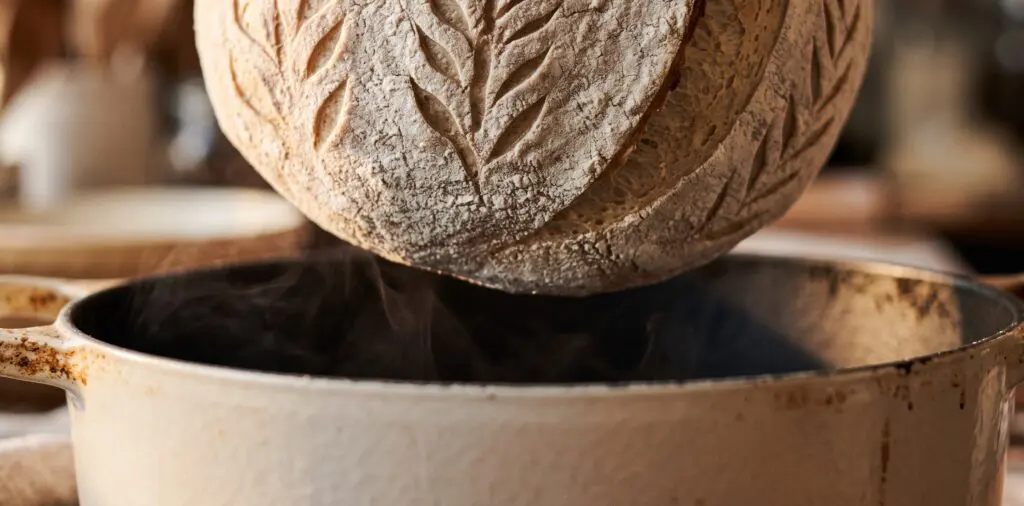 A flour-dusted sourdough boule with a scored pattern mid-drop into a preheated enameled Dutch oven.