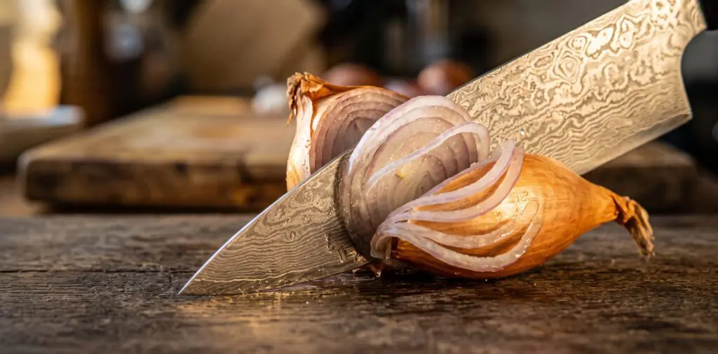 A Damascus chef's knife mid-slice through a golden shallot on an aged oak surface.