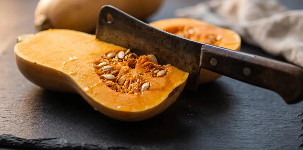 A butternut squash mid-split by a heavy cleaver, revealing deep amber flesh and pale cream seeds.