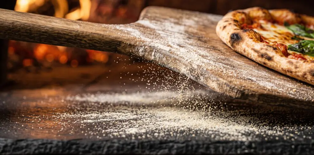 A freshly topped pizza mid-launch from a wooden peel onto a hot baking stone, semolina grains rolling beneath.