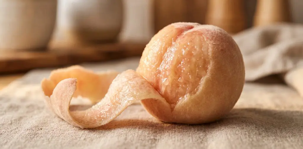 A blanched peach with its fuzzy skin peeling back in a single long sheet, vivid golden-pink flesh revealed beneath.