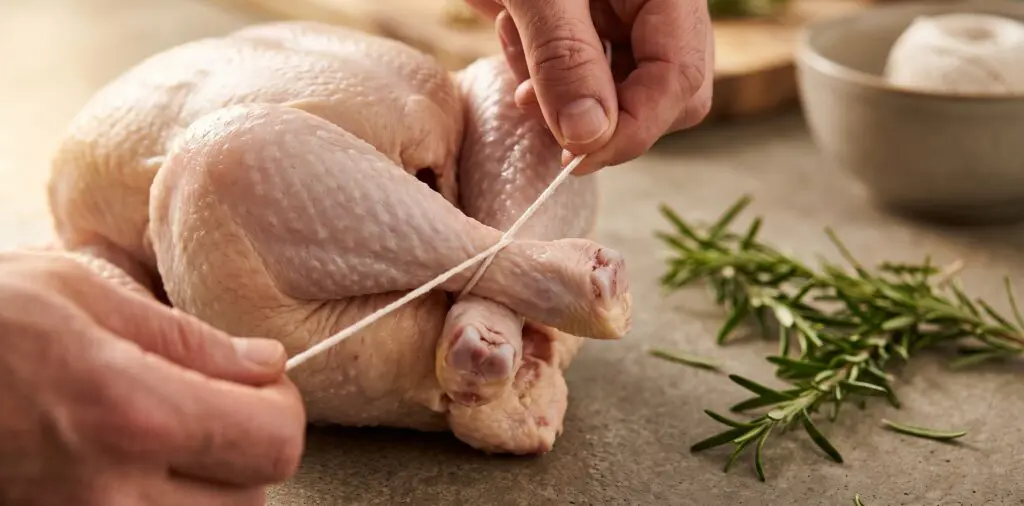 A whole raw chicken being trussed on a board, hands pulling kitchen twine to cross and secure the legs.