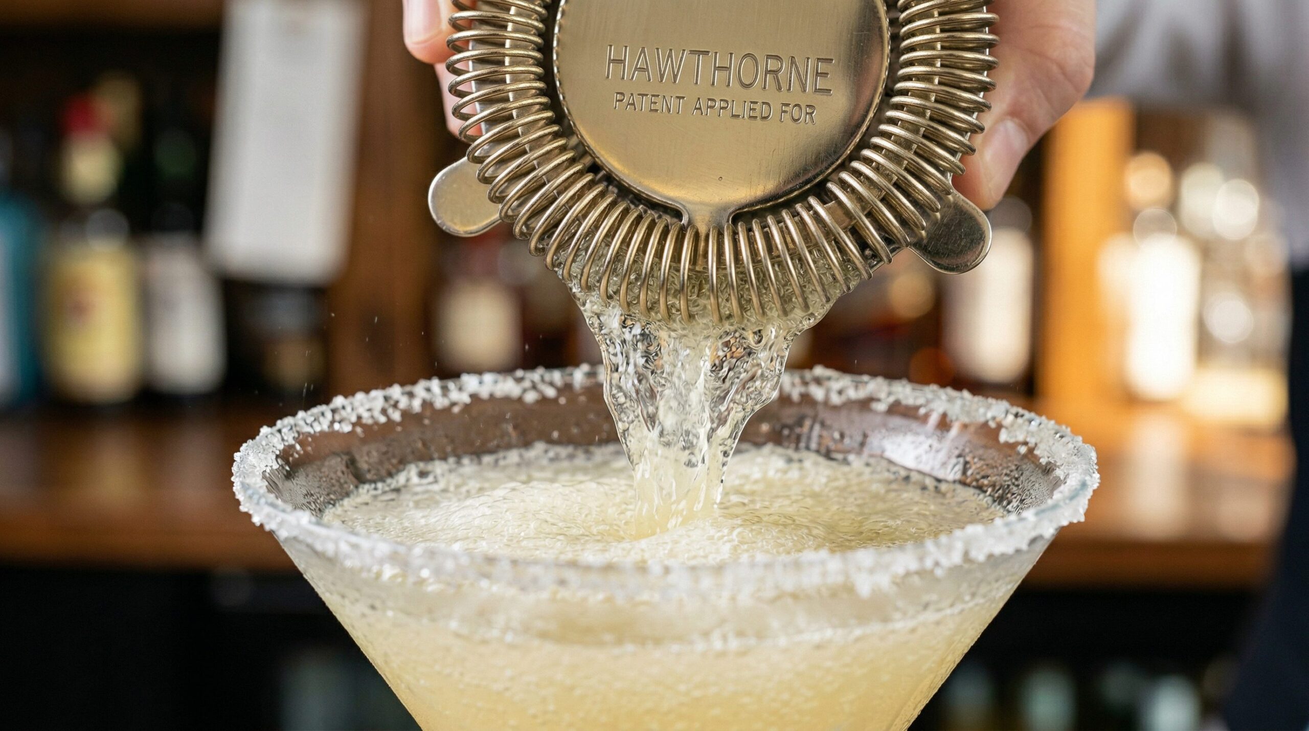 A frosty pale-yellow cocktail being strained through a Hawthorne strainer into a sugar-rimmed crystal martini glass
